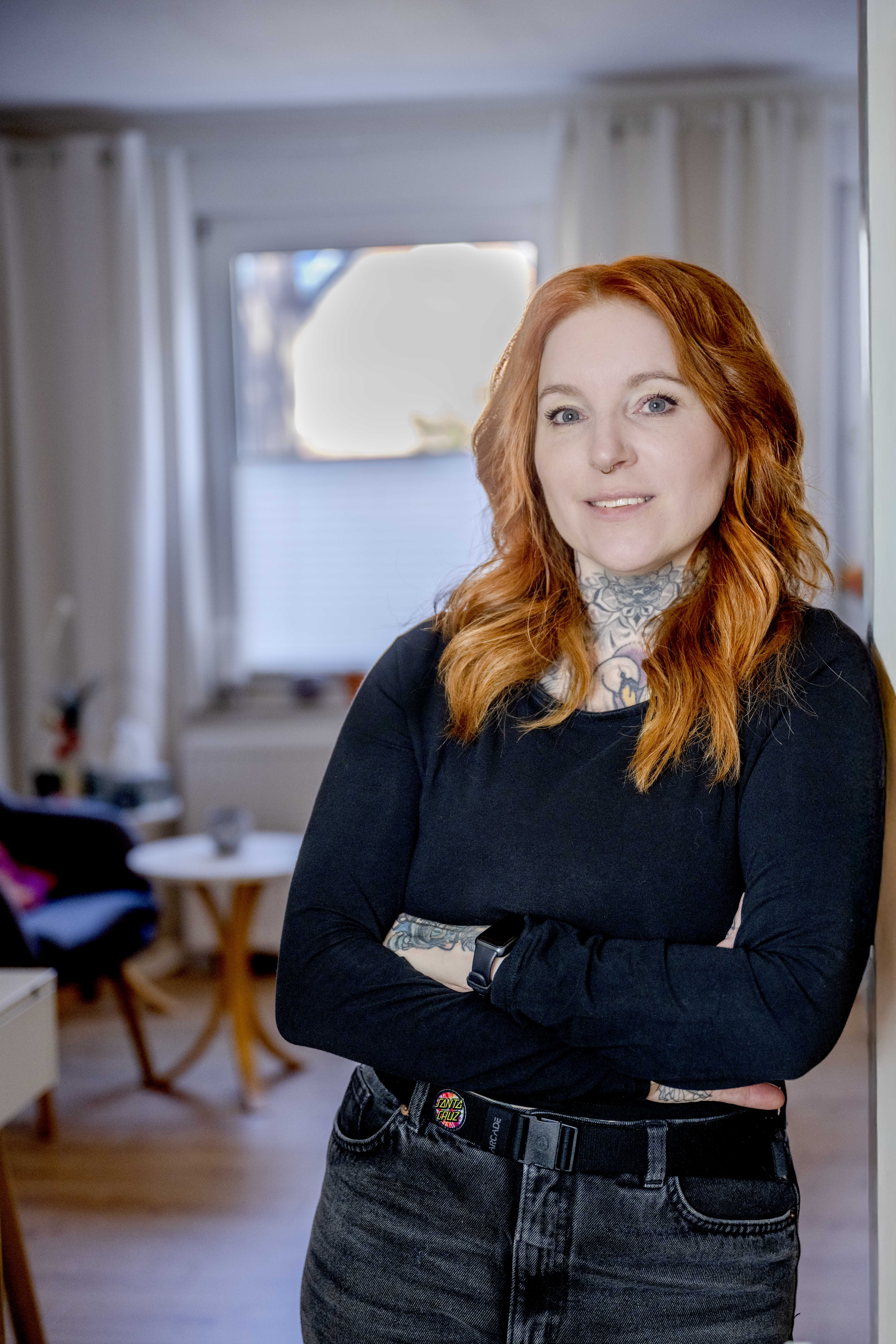 Startseite Manuela Becker, Manuela Becker, Rehabilitationspädagogin (BA) sowie systemische und psychosoziale Beraterin, mit kupferfarbenen Haaren und sichtbaren Tattoos, in schwarzem Oberteil, steht mit verschränkten Armen selbstbewusst und einladend lächelnd vor einem modernen Praxisraum.